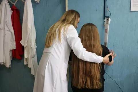 A female doctor with her back to camera helps a young girl on the scales