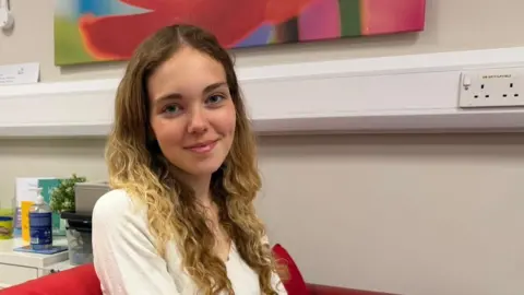 BBC Molly looks directly into the camera. She has curly blonde hair and dark blue eyes and is wearing a white blouse.