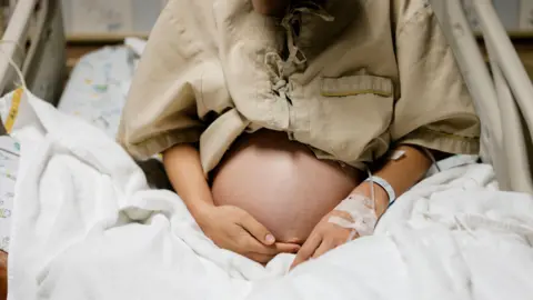 Getty Images Close-up of belly a heavily pregnant person in a hospital bed, with an IV attached to her arm.