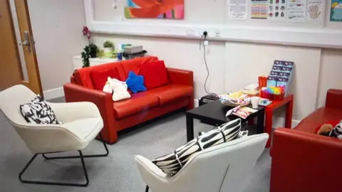 A room in the Nook centre. Two red leather sofas covered in red,white and blue scatter cushions face to face. Black and red tables are between them covered in leaflets. Two white chairs with black and white cushions in front of the sofas.