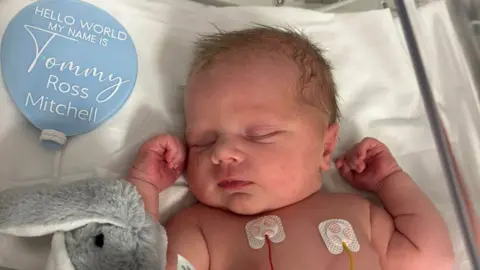 McGillivray Family Newborn baby Tommy in a cot. He has his hands up next to his head, his eyes closed and soft toy rabbit next to him. A blue balloon reads his name.