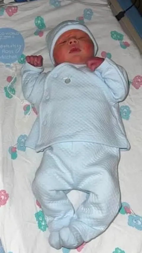 McGillivray Family Newborn baby Tommy in a cot. He has his hands up next to his head, his eyes closed and he is wearing a blue sleep suit.