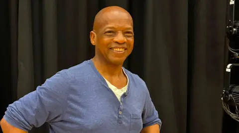 Hayden Etienne, who is wearing a blue long-sleeved T-shirt, is smiling at the camera and is standing in front of a black curtain.