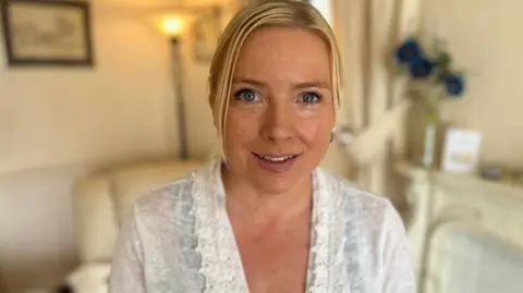 BBC A woman in a white top smiles at the camera. Behind her, out of focus, are a chair, lamp and flowers.
