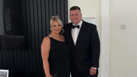 McGillivray Family Chelsea McGillivray wearing a black dress with her partner Jack who is in a black suit, white shirt and black bow tie. They are standing together, smiling at the camera.