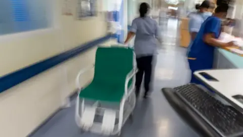 PA Media A nurse is moving a hospital chair along a corridor while two other nurses attend to paperwork