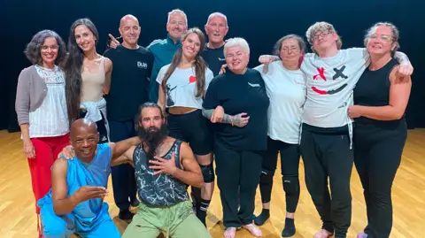 BBC A group of men and women, who are all wearing gym clothing, are gathered together in a dance studio for a photograph.