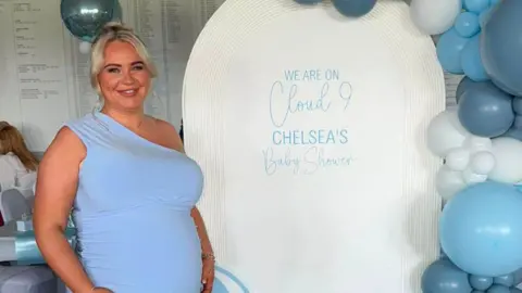 McGillivray family Chelsea McGillivray. She is wearing a blue one-shoulder dress and her blonde hair is tied into a bun. She is smiling at the camera.