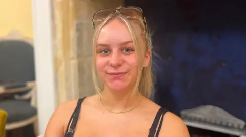 BBC Kelci Hill-Meldrum sits in a chair. She has long blonde hair tied up and a pair of sunglasses propped on the top of her head.