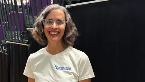 Claire Morris, who is wearing a white T-shirt with the words Fallen Angels Dance Theatre, is smiling at the camera