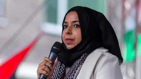Jo Holland/BBC Apsana Begum MP holding a microphone at a demonstration. She is wearing a black hijab and has a patterned black scarf with a cream coat. There is a building and out-of-focus flags behind her.