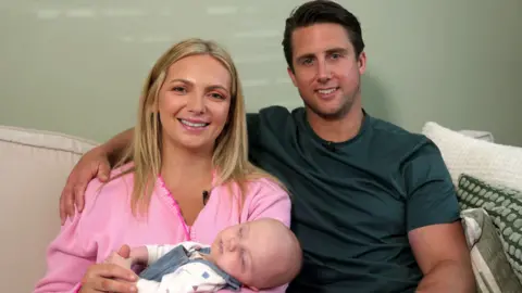Sleeping baby in the foreground, wearing a white long-sleeved top and blue dungarees, resting across the lap of a seated adult in a pink knitted cardigan. Another seated adult in a dark green short-sleeved top sits alongside with an arm resting behind the first adult. Both adults are seated on a light-coloured sofa with patterned cushions. Small clip-on microphones are attached to the adults’ clothing. A plain light-coloured wall forms the background.
