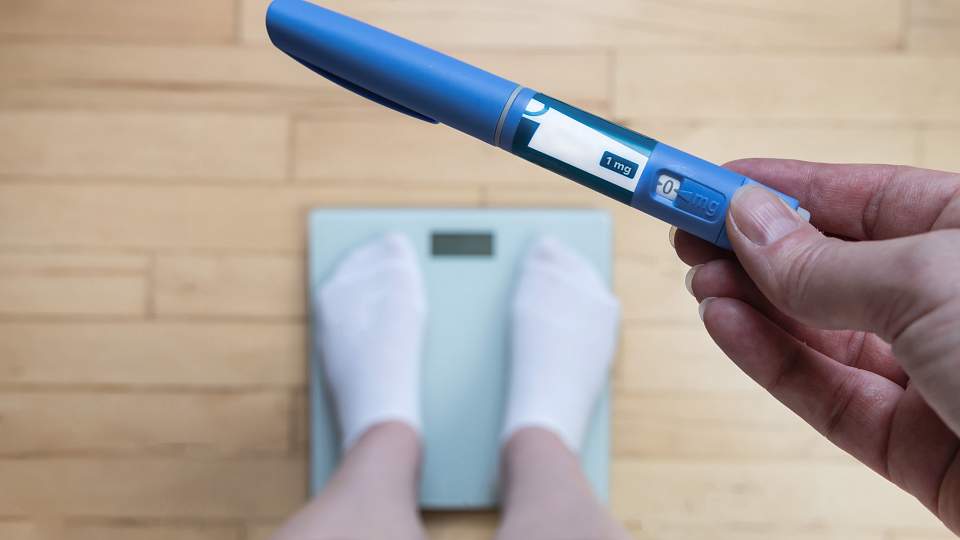Downward shot of a hand holding an Ozempic pen and feet standing on a scale.