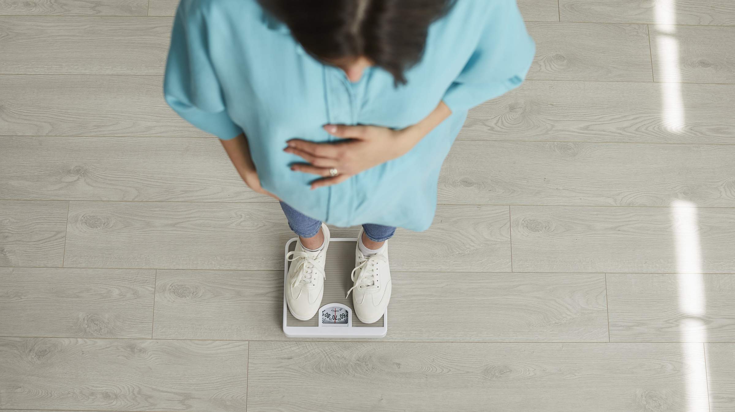 Overhead view of a person standing on a bathroom scale with hands resting on their abdomen, wearing a turquoise shirt and white sneakers on light wood floor