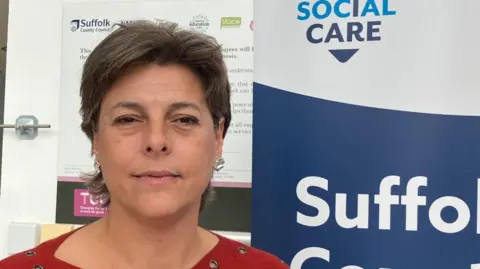Vikki Irwin/BBC A woman is standing against a notice board which has Suffolk County Council written on it. She is wearing a red top and has silver earrings.