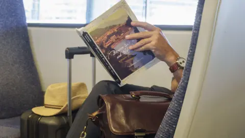 Getty Images A person is sitting on a train seat in a carriage. They are holding a large book and are sitting next to a brown leather bag and have a black suitcase in front of them.