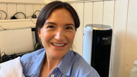 Vikki Irwin/BBC A woman is sitting on a wooden pew with white wood paneling in the background. She is wearing a blue shirt and has a towel over her shoulder. She has her hair tied back and is smiling at the camera.