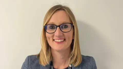 Contributed A woman is wearing a blue blazer and black T-shirt. She is standing against a plain cream backdrop. She is smiling and wearing blue rimmed glasses
