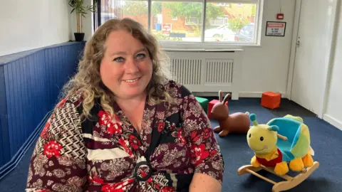 Vikki Irwin/BBC A woman is sitting in a room with blue carpet and blue wood paneling. She is wearing a multi-coloured top and is smiling at the camera. In the background you can see soft play toys.