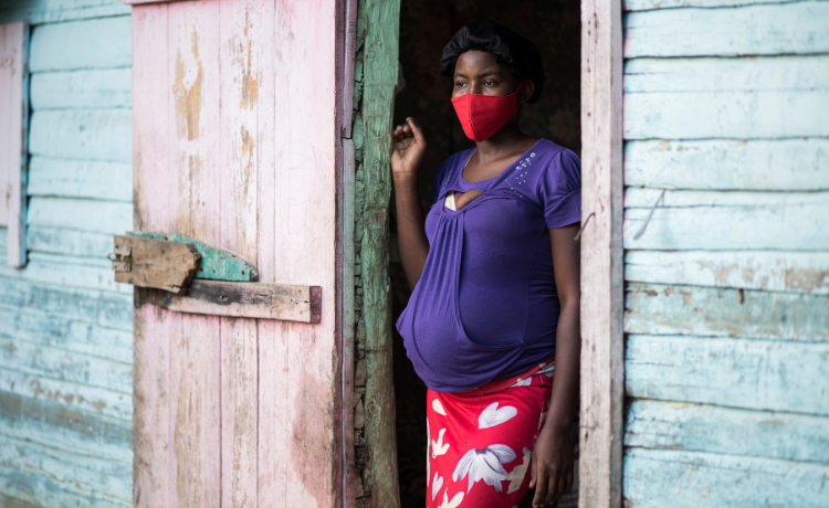 A pregnant woman in a dark purple top and red and white patterned skirt wears a red facemask while leaning against the pale pink doorframe of a turquoise-coloured wooden building