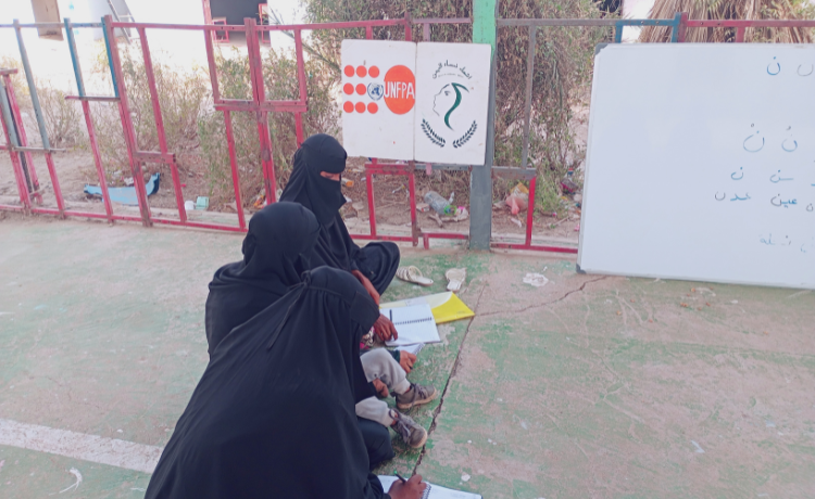 A row of women in black abayas and niqabs sit on the floor outside writing in textbooks; behind them a UNFPA sign is visible