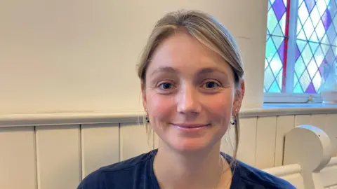 Vikki Irwin/BBC A woman is sitting on a wooden pew with a stained glass window in the background. She has her hair swept up in a ponytail and is smiling. She is wearing a blue T-shirt.