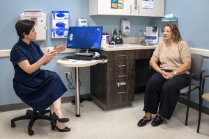 A physician, left, talks to a woman patient in a medical office. The physician gestures with her hands while seated next to a laptop and monitor.